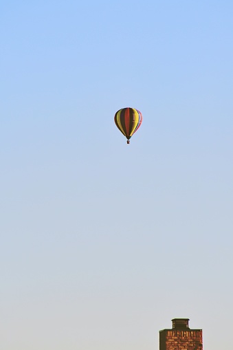 Up over the roof tops a hot air balloon soar against a blue sky showing off it’s canopy of bright colors