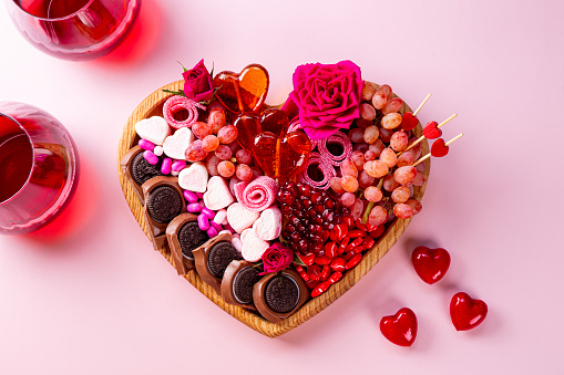 Snack board in shape of heart with berries, chocolate and sweets for Valentine's Day and two glasses of red wine on a pink background