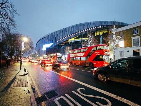 London, UK - 4 December, 2023: traffic and red London double decker buses on the High Road outside the Tottenham Hotspur stadium in north London, UK. It is early evening and the headlights of cars and vehicles are reflected on the wet surface of the road.