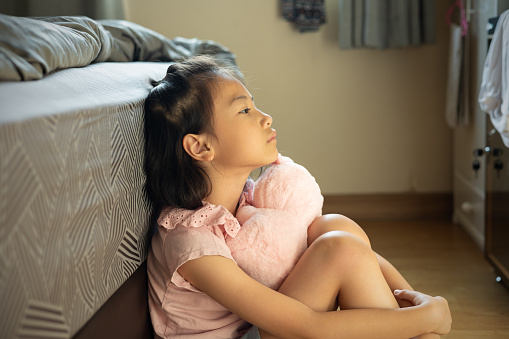 Adorable Asian kid girl is sitting on the bedroom floor alone. Her face thoughtfully expresses anxiety, sadness, sorrow and depression in her eyes and face with lonely and exhausted posture.