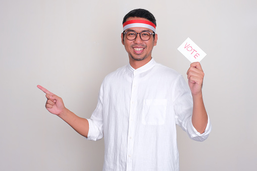 Indonesian man smiling and pointing to the right side while holding election ballot