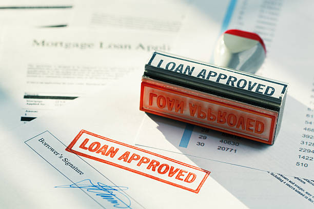 “LOAN APPROVED” Approval Red Rubber Stamp Approving Mortgage Application Document "Subject: Horizontal view of a rubber stamper with the words aLOAN APPROVEDaA resting on a pile of loan papers, including a borroweraas signature page stamped in red, showing approval was granted. Bright lighting and long shadows create a positive mood." types of loans stock pictures, royalty-free photos & images