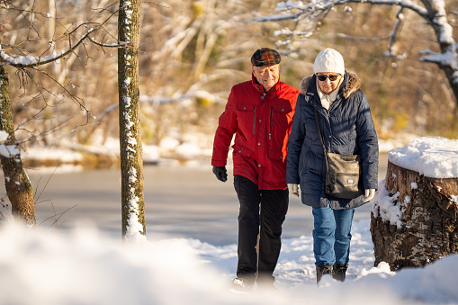 senior couple dressed in warm winter jackets on walk through natural park with lake outdoors on sunny cold day and much snow
