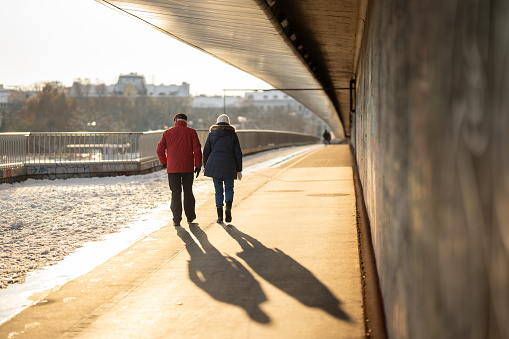 rear view of senior couple in warm winter jackets walking over footbridge for pedestrians and cyclist over river in city on cold sunny winter day, shadows of people on the ground