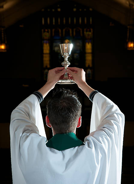Priest in Robe Blessing Wine for Communion Back of priest raising chalice toward heaven. Focus on priest with back lighting illuminating his hair and robe. anglican stock pictures, royalty-free photos & images