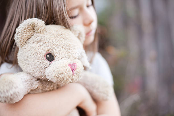 Color photo of a sad, lonely 6-7 year old girl hugging an old, raggedy teddy bear outside an old, abandoned house.