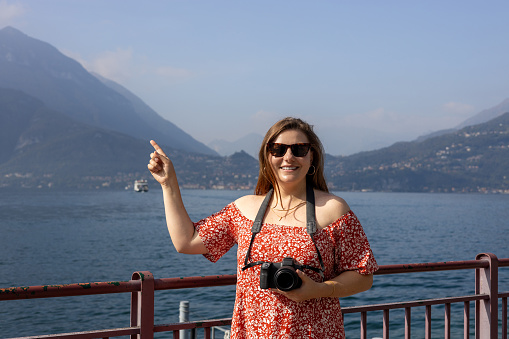 Happy brunette 30s woman enjoying summer holidays. Concept of vacation and travelling. Varenna, Italy. Lake Como. Redhead girl pointing finger and holding a camera outdoors