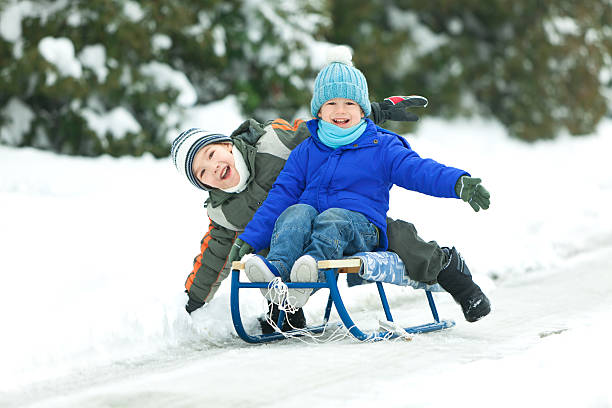 Two smiling little boys sledding in snow through winter landscape. Some visible motion blur.