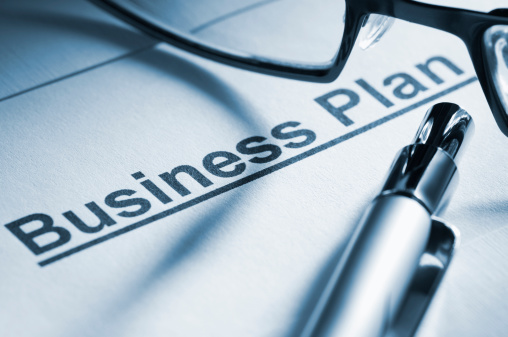 A professional business plan lying under a pair of reading glasses and a ballpoint pen. The document and glasses are backlit and cast a strong shadow across the business plan. The focus is selective and sharp on the word Business. The image has been toned blue.
