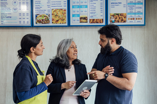 Waist up of a mature female business owner and her son working in their family run pizza shop in North Yorkshire, England. They are wearing navy shirts and aprons, they are with a mature female financial advisor, she is smartly dressed and holding a digital tablet.