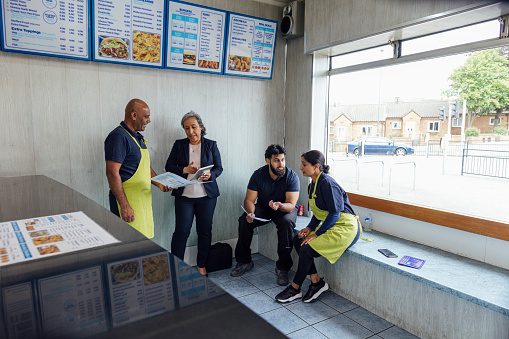 Over the counter view of a mature couple and their son working in their family run pizza shop in North Yorkshire, England. They are wearing navy shirts and aprons, they are with a mature female financial advisor, she is smartly dressed and holding a digital tablet.
