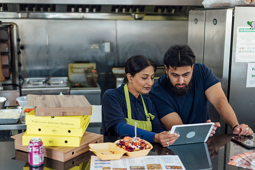 Close up of a mature female business owner and her son working in their family run pizza shop in North Yorkshire, England. They are wearing navy shirts and the woman is wearing an apron. They are brainstorming business ideas and using a digital tablet at the checkout counter.