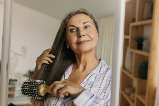 Beautiful, charming elderly female combing grey hair with round hairbrush standing against shelves in hallway or bedroom in front of mirror, looking at her reflection, getting ready to go to bed