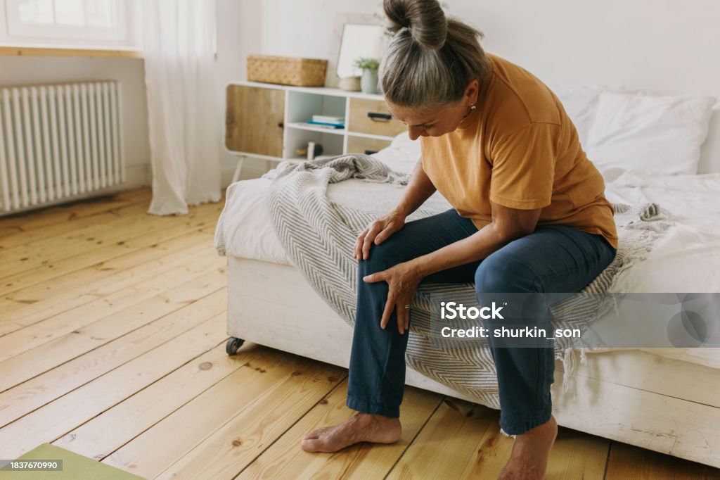 Elderly woman have pain in knee. Mature female suffer with trick knee sitting on bed at home, cozy bedroom interior. Growing old, take care of your health, health problems concept Pain Stock Photo Elderly woman have pain in knee. Mature female suffer with trick knee sitting on bed at home, cozy bedroom interior. Growing old, take care of your health, health problems concept Pain Stock Photo