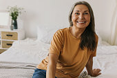 Sweet, adorable modern grandmother in casual T-shirt and jeans laughing sitting on bed chilling after home tasks, feeling joy and happiness, relaxing in bedroom while pie are being cooked in oven