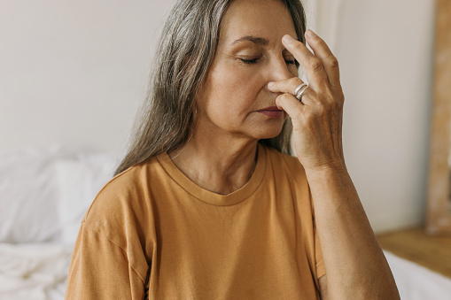 Crop shot portrait of mature grey-haired female in orange T-shirt doing breathing techniques, holding nose, inhaling and exhaling, sitting with closed eyes against white background. Healthy lifestyle