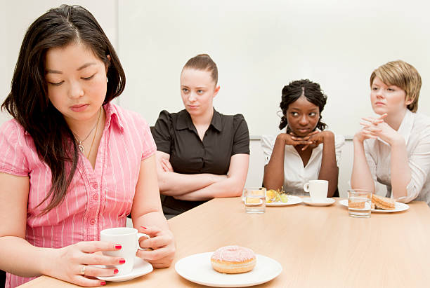 Workplace Exclusion A young woman sits alone while three of her colleagues look at her with disdain.  Shallow depth of field - focus on the excluded girl in the foreground.Click exclusion group of people separation fish out of water stock pictures, royalty-free photos & images