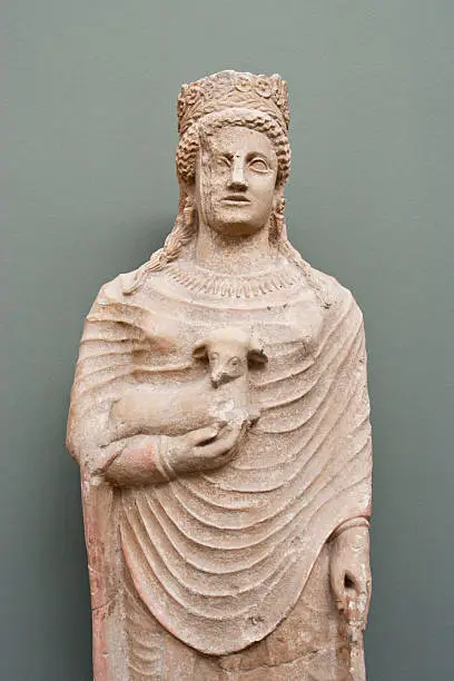 "Statue of a female votary on gray background. Cyprus, 5th - 4th cent. BC. Limestone.The woman wears a wreath of laurel and flowers. In her hand she holds a small calf, which she presents to a divinity, probably Aphrodite. Placed as a votive in a sanctuary.Ancient art photographed in Carlsberg Glyptotek, Copenhagen, Denmark."