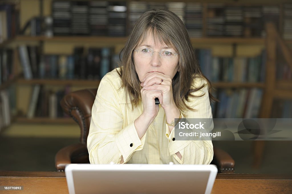 Serious businesswoman with laptop "Mature businesswoman looking very serious as she sits at her her desk in front of her laptop, in a traditional book-lined office/study settingMore like this" In Front Of Stock Photo