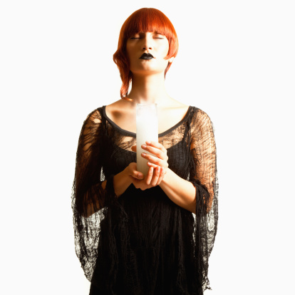 A beautiful young woman in Gothic avant-gard attire holds a white candle and meditates with eyes closed. Her hair style is fashion forward and dyed red. Square format and white background.