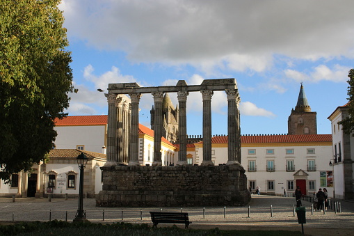 The roman temple of Augustus seen in Jardim Diana in the city of Évora (Alentejo, Portugal). Behind it it's possible to notice other buildings from the city of Évora. Sky is light blue with some clouds. There is a tree in the left side and a light pole (turned off). Orange of the ceilings call attention.