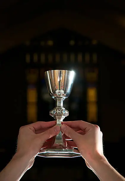 Hands holding silver chalice in dark church. Blurred stained glass in back ground.
