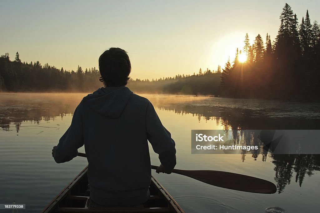 Sunrise Paddle A young man paddles a canoe on a glassy calm lake in the early morning. Canoeing Stock Photo