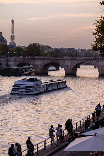 Paris, France - October 8, 2023 : View of people walking next to the river Seine, a touristic boat and the Eiffel Tower in the background in Paris France at sunset