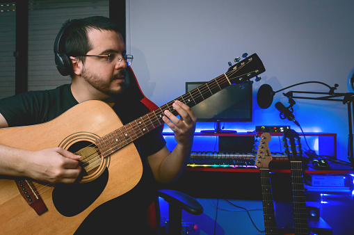 portrait of latin male classical music guitarist, with beard, glasses and headphones sitting in his home studio playing the guitar, checking the sound and tuning before recording.