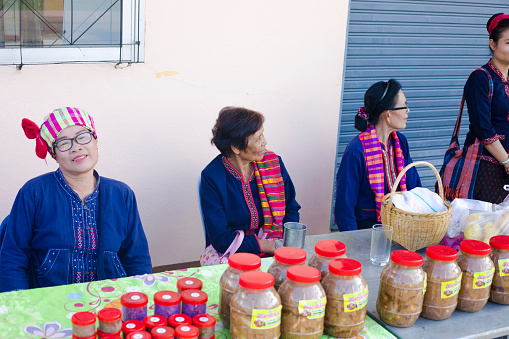 Mature and senior thai women are sitting at table with local homemade thai food in packages. Scene is at a small local food market event in Chiang Khong in Chiang Rai province. Three women are sitting behind a tabke in front of a house.  A fourth woman is standing close by. Person is talking to two sitting women.On tabke are bottles with food.  One mature woman is wearing glasses. Three women are wearing traditional blue farm fashion and thai paccama scarf