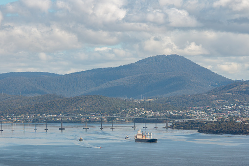 Hobart, Tasmania, Australia - December 12 2022: Derwent Estuary with cargo vessel and tug boarts cruising past Derwent bridge towards entrance of harbour