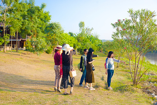 Group of adult thai people at waterfront of Mekong river at late afternoon. Scene is in Chiang Khong area in Chiang Rai province in north Thailand. People are standing close to river in landscape and are watching into scenery. There are mostly women dressed in different styles. Two are gesturing with hands into direction and one is wearing a straw hat. People are watching over river towards Laos
