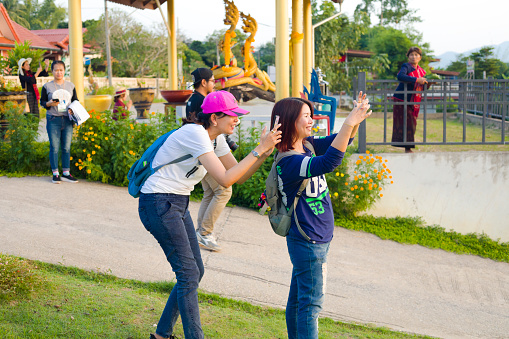 Two auldt thai women are taking photos with mobile phones at waterfront of Mekong river in Chiang Khong area in Chiang Rai province in north Thailand. One woman is wearing a pink colored cap. In background are more thai people. People are visiting local Nakhon shrine at river. Women are photographing river and geographical border with landscape to Laos.