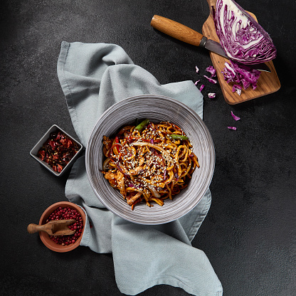 Top view, square frame, featuring udon noodles stir-fried with chicken and vegetables in garlic sauce. Chopsticks and textile elements accentuate the dish against a dark, dramatic backdrop.