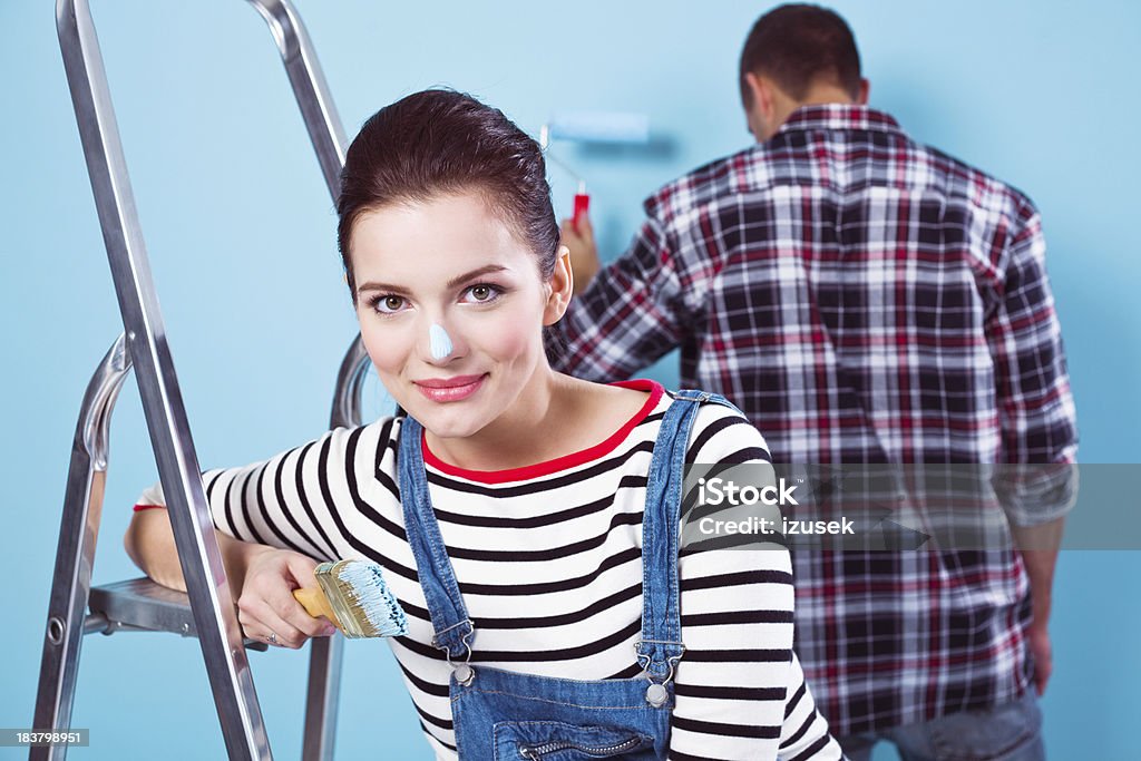 Beautiful female house painter "A young adult couple painting the blue wall. Focus on the beautiful brunette with the paintbrush and painted nose, standing next to the ladder in the foreground, looking at camera and smiling. Her partner in the background with paint roller." 20-24 Years Stock Photo