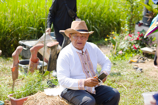 Thai senior man with cowboy hat and traditional clothes and white shirt is sitting on hay near a rice paddy. Scene is at field area with other agriculture and river crabs farming and a small vegetable market by local farmers  in rural lanscape in Chiang Rai province. Man is holding a mobile phone in his hand. In background is another person