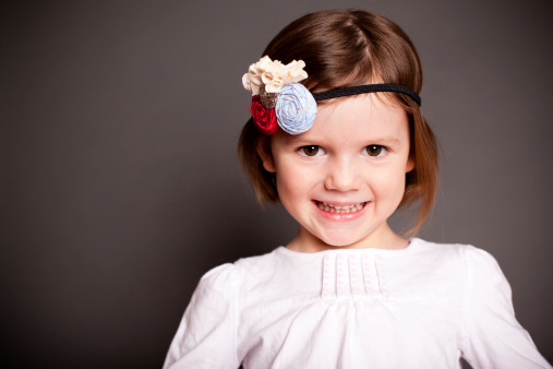 Color image of a pretty little girl smiling at the camera while wearing a pretty headband and on gray background.