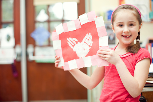 A smiling student girl stands in her class and shows off her valentines day card she made out of red, white, and pink paper and hand prints.  Horizontal with copy space.