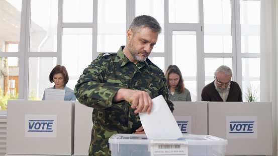A military man, a soldier, putting a voting ballot into the box at the polling place during elections