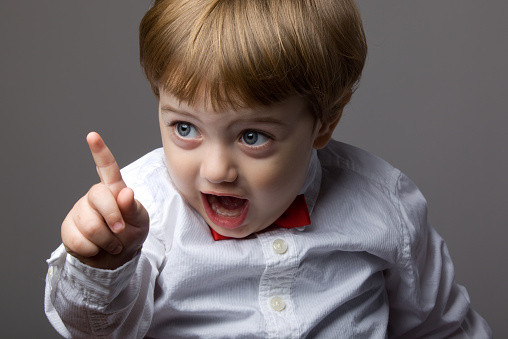 Serious Teen boy with raised index finger, looks at camera, stands over grey background, wearing in casual clothes. Expressive facial expressions