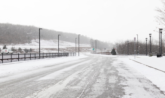 This remote highway rest stop parking lot is sloppy and slushy. Heavy, wet snow is falling along Route 81 in central New York state during a winter blizzard. A couple of cars can be seen passing by the entrance ramp up to the main road, but very few people are traveling in this weather.