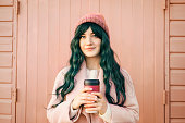 Stylish young smiling hipster woman with color hair wearing trendy peach color coat and hat with reusable coffee cup on wooden background. Urban seasonal city street fashion. Color of the 2024 year.