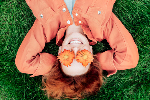 Top view happy smiling redhead Woman in trendy peach color jacket and gerbera Flower glasses lying on green grass. Positive Emotion people. Color of the 2024 year. Spring, summer mood.
