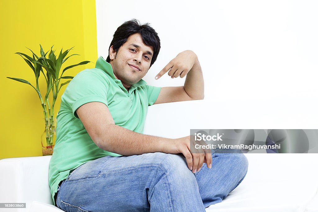 Jovem alegre indiana bonito macho adulto sentado em um sofá - Foto de stock de Pointing At Self royalty-free