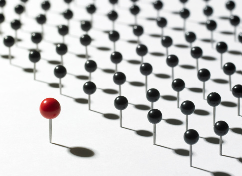 A single large red map pin standing up in front of several rows of smaller black pins. Lighting and focus is sharp on the red pin. Excellent for educational concepts.