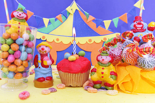 A studio shot of birthday party decoration with circus tent and clowns. Front view of cupcake with a burning candle, clowns candles, vases with lollipops and candies on the foreground. A colourful circus tent with garlands stands at the background. 