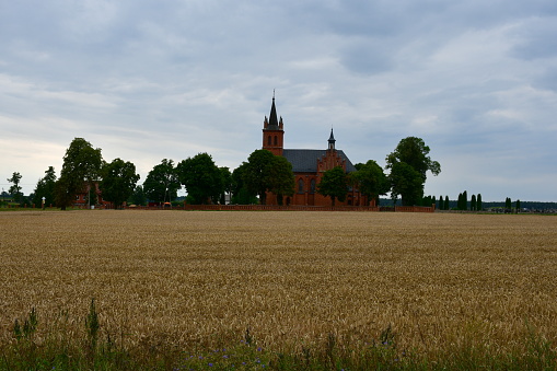 A view of a church or chapel made out of red brick surrounded from all sides with small trees and shrubs situated in a close proximity to vast field, meadow, or pastureland full of crops