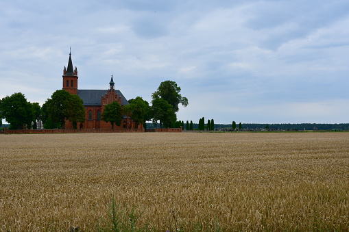A view of a church or chapel made out of red brick surrounded from all sides with small trees and shrubs situated in a close proximity to vast field, meadow, or pastureland full of crops