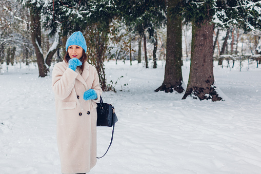 Elegant young woman walking in snowy winter park wearing white sheepskin fur coat blue knitted hat holding handbag under falling snow. Warm stylish clothes