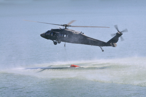 horizontal image of a black hawk helicopter filling a bambi bucket in a body of water. helicopter is slightly obscured with water vapor.